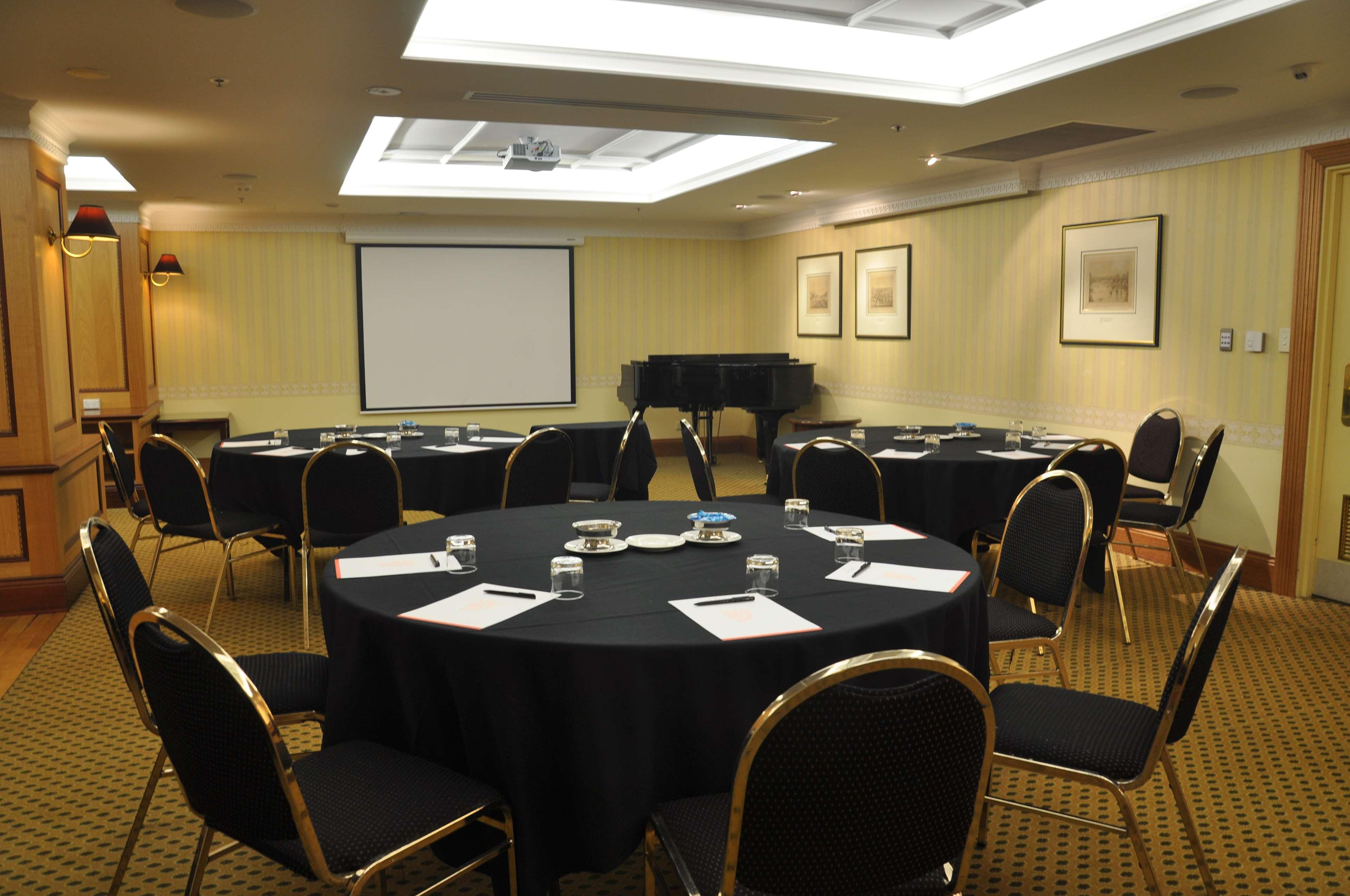 Stamford Plaza Adelaide Otel Dış mekan fotoğraf The photo shows a conference room set up for a meeting or presentation. There are several round tables covered with black tablecloths, each surrounded by black upholstered chairs. On the tables, there are notepads and glasses of water, indicating tha