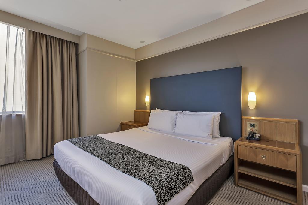 Stamford Plaza Adelaide Otel Dış mekan fotoğraf The photo shows a modern hotel room. It features a double bed with a white duvet and a decorative grey and black throw. The headboard is a solid dark color. There are two bedside tables on either side of the bed, each with a lamp. Natural light enter